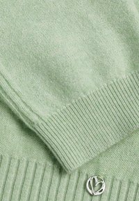 Tissu tricoté vert clair doux avec des bords côtelés et un petit ornement en métal argenté en forme de cœur rond.