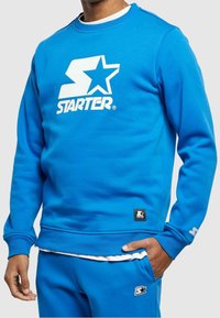 Felpa blu con un grande logo bianco a forma di "S" e stella, polsini e orlo a coste, texture liscia, abbinata a pantaloni coordinati.