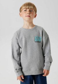 Sudadera gris con cuello redondo, puños acanalados y un gráfico en color teal que dice "Keep Going" en el pecho, combinada con jeans azul oscuro.