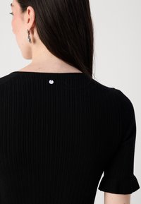 A woman with long, dark hair wearing a black ribbed top, showing a small decorative button at the back and short, frilled sleeves.