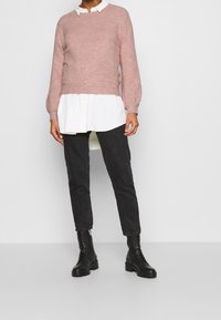 Jasnoróżowy sweter z okrągłym dekoltem, noszony na białej koszuli z kołnierzykiem. Czarnie obcisłe jeansy i czarne, sznurowane botki do kostek dopełniają stylizację.