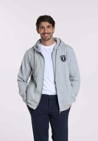 Hoodie gris zippé avec cordons de serrage et poche avant, arborant un logo en forme de bouclier noir. Porté sur un t-shirt blanc, associé à un pantalon bleu marine.
