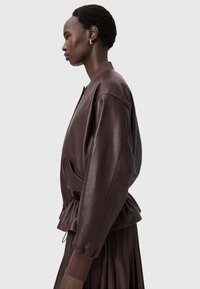 Burgundská kožená bomber bunda s uvolněným střihem, elastickým lemem a žebrovanými manžetami. Obsahuje postranní kapsy a hladký povrch.