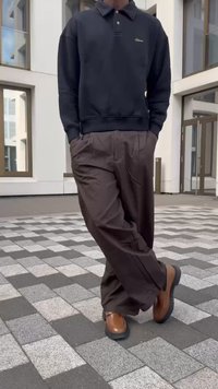 Sweatshirt preto com um logótipo subtil, calças castanhas escuras com bolsos e sapatos castanhos. O conjunto combina elementos casuais e descontraídos.