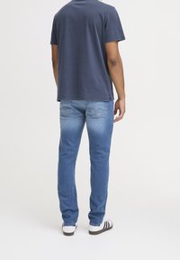 Man wearing blue faded jeans, navy blue short-sleeve shirt, and white sneakers standing facing away on plain white background.