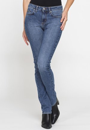 Donna che indossa jeans slim blu, top nero e stivaletti neri, in piedi con le gambe incrociate davanti a uno sfondo semplice.