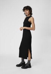 Vestido negro sin mangas de canalé con aberturas laterales. Presenta un escote alto y un diseño ajustado, combinado con zapatos gruesos negros y calcetines grises.