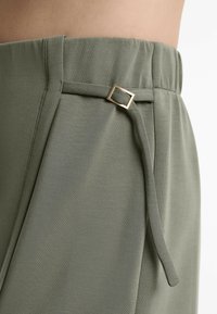 Pantalons vert olive à coupe décontractée, avec une boucle de ceinture en métal doré en forme de carré et un tissu doux et texturé. Taille élastique pour plus de confort.