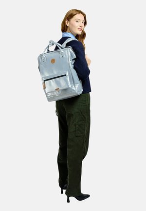 Frau trägt einen dunklen Pullover und grüne Hose, trägt einen glänzenden hellblauen Rucksack, steht und schaut über ihre Schulter zurück.