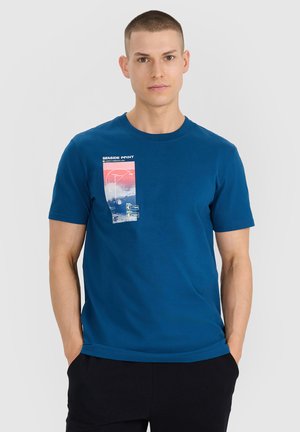 Młody mężczyzna z krótkimi włosami, ubrany w niebieski t-shirt z nadrukiem nadmorskiego krajobrazu, stoi z rękami w kieszeniach czarnych spodni.