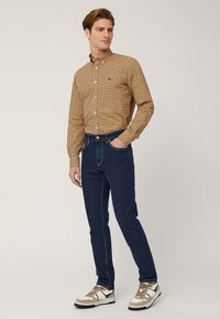 Camicia a maniche lunghe con un motivo a quadri marrone, giallo e bianco; abbinata a jeans slim-fit blu scuro e sneakers bianche con dettagli beige.