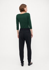 Grønn tettsittende topp med trekvart ermer, kombinert med svarte bukser med elastisk liv. Flate sko fullfører antrekket. Vist fra baksiden.
