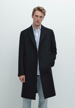 Ung mand iført en lang sort frakke over en hvid skjorte og lyseblå jeans, stående med den ene hånd i lommen mod en ensfarvet baggrund.