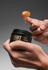 STMT Fiber Pomade i en svart burk med guldfärgad text, hålls i en hand. En trögflytande, bärnstensfärgad substans lyfts från burken.