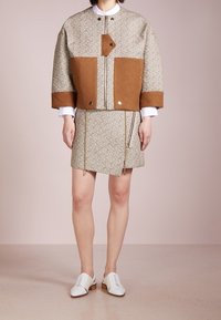 Chaqueta beige texturizada con detalles marrones y cierre de snap, combinada con una falda a juego y zapatos blancos con cordones, que presentan un corte moderno.