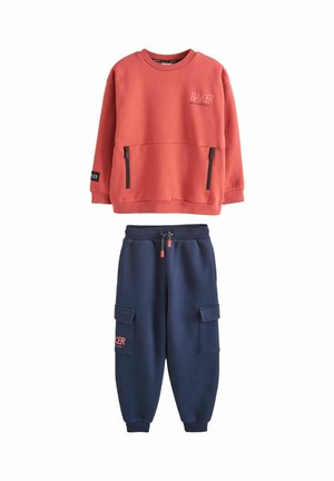 Red long-sleeve sweatshirt with black zippers and navy blue cargo joggers with drawstring waist and side pockets.