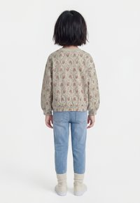 Bērns stāvot muguru pagriezies, valkājot ziedu raksta džemperi, gaiši zilas džinsas, krēmkrāsas zeķes un bēšas sporta kurpes uz vienkārša fona.