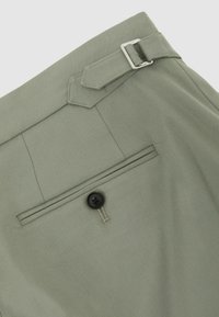 Bolsillo trasero de pantalones verde claro con un solo botón negro y correa lateral ajustable con hebilla plateada.