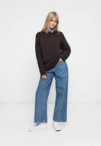 Kvinne stående mot en hvit vegg, iført en mørk brun genser over en blåstripete skjorte, vide jeans og hvite platå-sneakers.