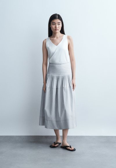 Jeune femme debout contre un mur uni, portant un haut blanc sans manches, une jupe midi rayée et des tongs noires, regardant légèrement sur le côté.