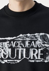 Svart sweatshirt med en riven grafikdesign och vit text som lyder "VERSACE JEANS COUTURE." Tyget är slätt med en ribbad halsringning.