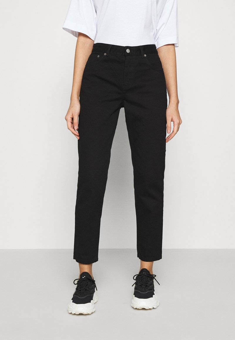 Personne portant un pantalon noir taille haute à jambes droites, une chemise blanche et des baskets noires et blanches, debout sur un sol gris clair.