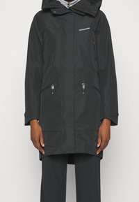 Cappotto impermeabile nero con cappuccio, cerniere sulle tasche frontali e tessuto testurizzato. Presenta una vestibilità comoda e maniche lunghe.