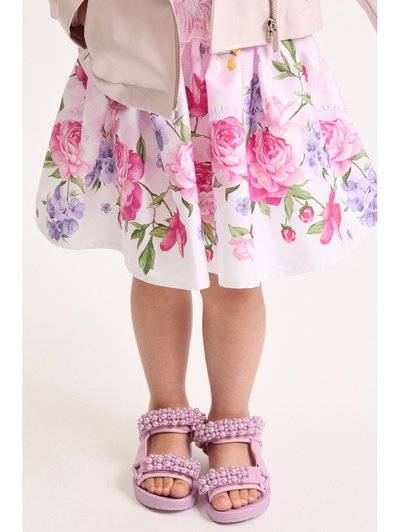 Παιδί που φοράει ρουμπινί φόρεμα με λουλουδάτο σχέδιο, ανοιχτό ροζ μπουφάν και ροζ σανδάλια διακοσμημένα με μαργαριταρένιες χάντρες.