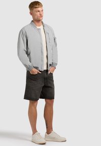 Jeune homme aux cheveux courts portant une veste bomber gris clair, un t-shirt écru, un short en denim noir et des baskets blanches, debout les mains dans les poches.