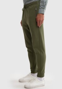 Oliven grønne sweatpants lavet af et glat stof, med en snøre i taljen, sidelommer med lynlås og ribbede manchetter. Båret med hvide sneakers.