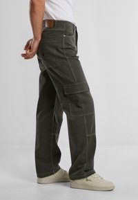 Pantalones cargo de algodón verde oscuro con costuras visibles, bolsillos laterales y un parche de cuero en la parte posterior. Combinados con zapatillas en color blanco roto.