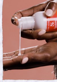 Vit flaska med röd etikett som dispenserar vit lotion i en hand. Handen har ljusa naglar, slät hud och en neutral bakgrund.