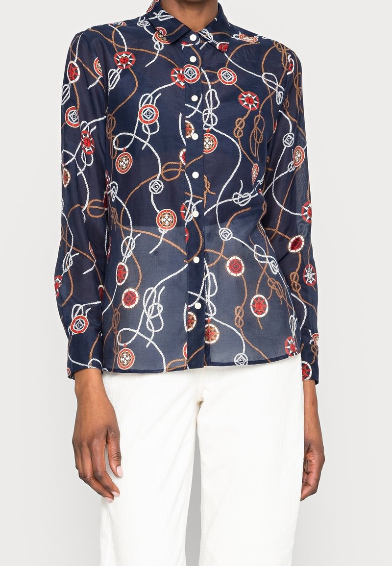 Chemise bleu marine à boutons avec manches longues comprenant des motifs de cordage nautique rouge, blanc et marron ainsi qu’un compas, portée avec un pantalon blanc.