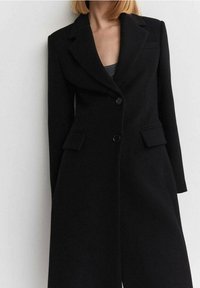 Donna che indossa un lungo cappotto nero su misura con rever a punta e due tasche anteriori a patta, in piedi contro uno sfondo chiaro e semplice.