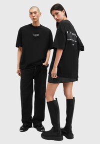 Svarte oversized t-skjorter på to modeller, med logo trykk. Den mannlige modellen kombinerer med mørke jeans; den kvinnelige modellen har på seg en kjole og knelengde støvler.
