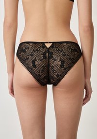 Zwart kanten lingerie met bloemmotieven, ingewikkelde details en gegolfde randen. Heeft een kleine sleutelgat uitsparing aan de achterkant.