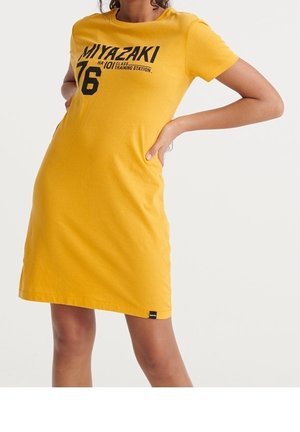 Woman wearing a yellow short-sleeve dress with black text "MIYAZAKI 76 Training Station" standing with hands on hips.