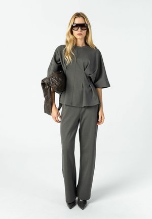 Vrouw draagt een oversized donkergrijze top en bijpassende broek, houdt een donkerbruine leren jas vast en draagt grote zwarte zonnebrillen.
