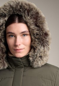 Olivovo zelená prešívaná bunda s veľkou kapucňou z umelého kožušinového materiálu v hnedých odtieňoch, s predsadeným zipsom a prešitými detailmi na látke.