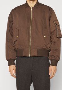 Brown bomber jacket with a zip front, ribbed cuffs and hem, and side pocket. Features a textured patch pocket on the sleeve.