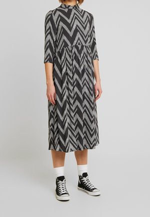 Osoba ubrana w sukienkę w czarno-biały zygzakowaty wzór z rękawami do połowy przedramienia oraz czarne wysokie trampki z białymi skarpetkami.