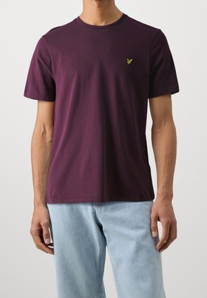 Mand iført en ensfarvet mørkelilla t-shirt med korte ærmer med en lille gul broderet logo på brystet og lyseblå jeans.