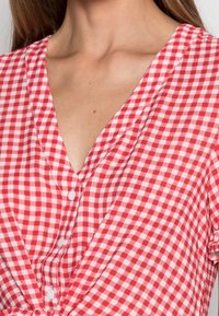 Chemise à motif vichy rouge et blanc avec un décolleté en V, des manches courtes et des détails de boutons sur le devant. Tissu en coton léger.