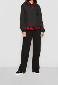 Person klädd i en svart zip-up jacka över en röd och svart rutmönstrad skjorta, med svarta cargobyxor och svarta sneakers med vita sulor.