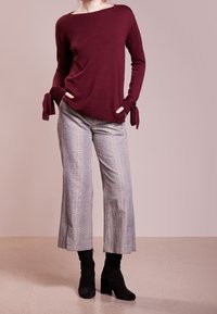 Maglione bordeaux con dettagli a cravatta alle maniche, abbinato a pantaloni a gamba larga grigio chiaro a quadri e stivaletti neri.