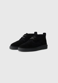 Stivali alla caviglia in suede nero con punta arrotondata, suola in gomma nera texturizzata e lacci neri. Design minimalista con dettagli sottili.