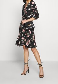 Schwarzes Blumenkleid mit pinken Blumen, dreiviertel Ärmel und A-Linien-Rock. Kombiniert mit schwarzen Riemchen-Heels und einer strukturierten schwarzen Handtasche.
