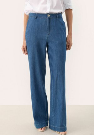 Personne portant un jean en denim bleu taille haute à jambes larges avec une fermeture par bouton et zip, associé à une chemise blanche et des sandales à bouts ouverts.