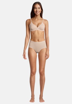 Frau steht barfuß und trägt einen beigen passenden BH und hochtaillierte Slips, lächelt vor einem schlichten weißen Hintergrund.