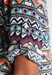 Mönstrad skjorta i flera färger med geometriska mönster i marinblått, turkos, rött och vitt. Metallknappsstängning vid manschetten. Material av bomull.